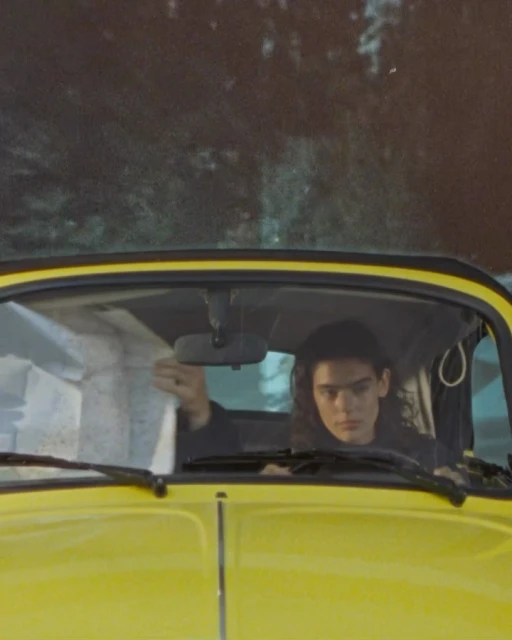 A young adult with long curly hair is seen in a yellow vintage car, looking at a map. The background shows a blurred landscape.