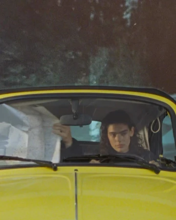 A young adult with long curly hair is seen in a yellow vintage car, looking at a map. The background shows a blurred landscape.
