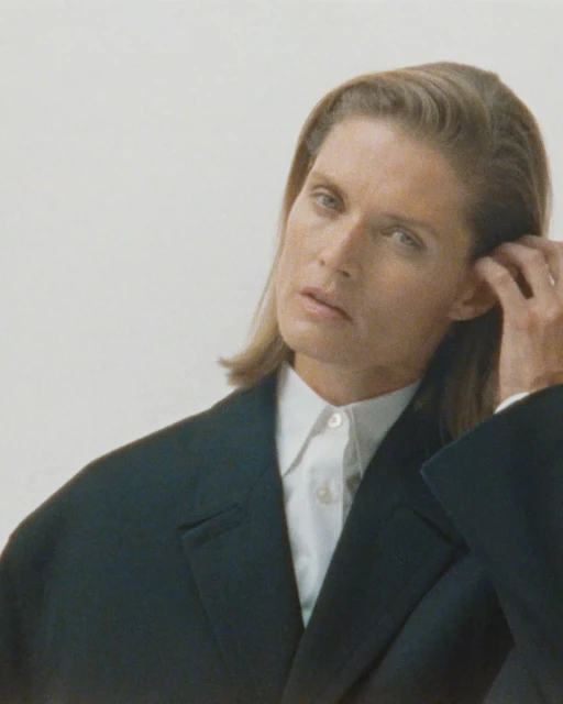 Portrait of a blonde woman in a dark blazer and white collared shirt, gazing with a slight frown against a plain background.