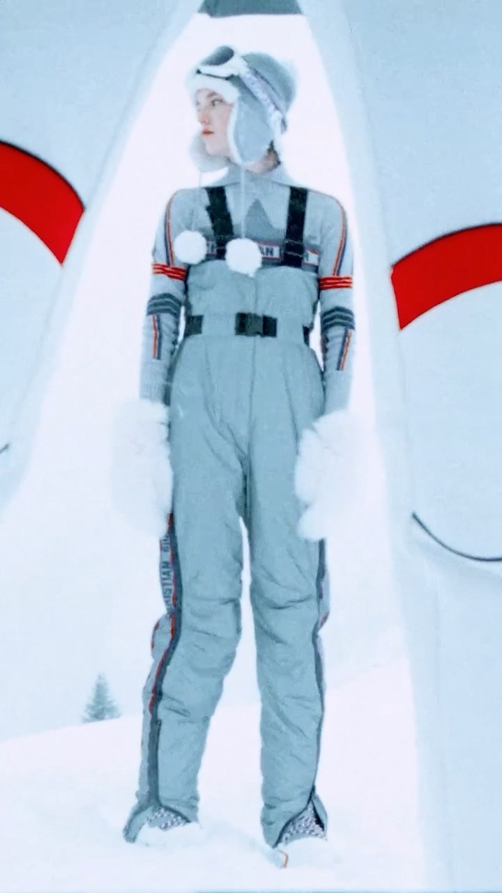 A woman in a grey snowsuit and a fur-trimmed hat stands in a snowy landscape with architectural features and a pine tree in the distance.