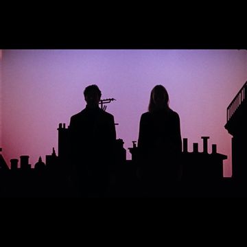 Silhouette of a couple standing on a rooftop at dusk, with city skyline and soft pink sky.