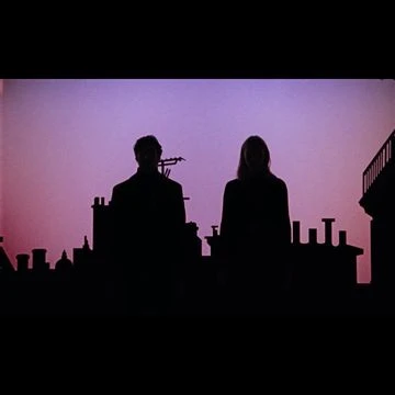 Silhouette of a couple standing on a rooftop at dusk, with city skyline and soft pink sky.