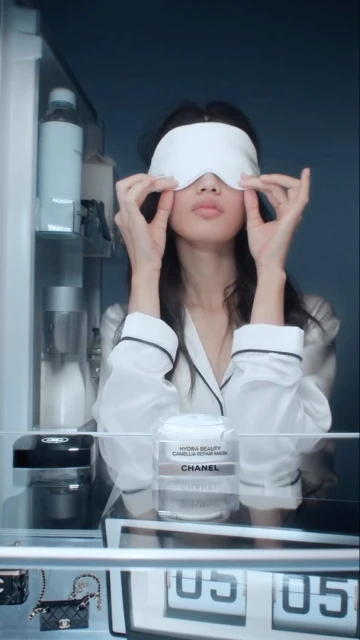 Adult female in pajamas wearing a sleep mask in front of an open refrigerator with beauty products, handbags, and a digital display.