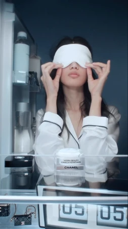 Adult female in pajamas wearing a sleep mask in front of an open refrigerator with beauty products, handbags, and a digital display.