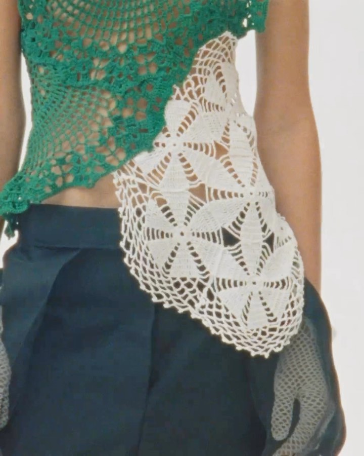 Cropped view of a person wearing a green and white crocheted top and dark blue shorts against a plain white backdrop.