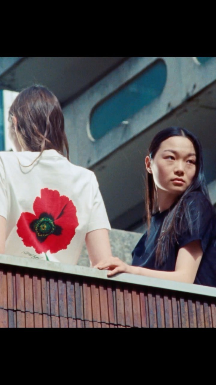 Two women pose outdoors with architectural backdrop. One wears a white t-shirt with a red poppy print; the other wears a dark top.