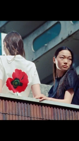 Two women pose outdoors with architectural backdrop. One wears a white t-shirt with a red poppy print; the other wears a dark top.