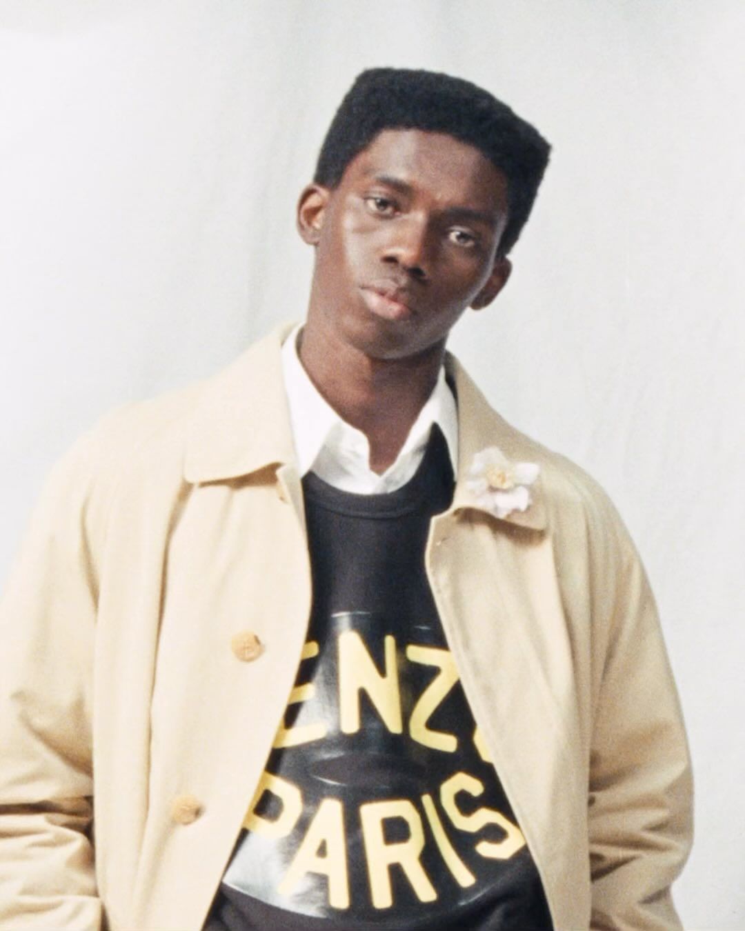 A young African-American man poses in front of a white backdrop, wearing a tan trench coat and a black Kenzo Paris sweatshirt.