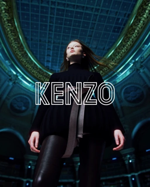 Fashion shot: Woman in black top and shiny black leggings poses in ornate, domed building under dramatic blue lighting and 'KENZO' text.