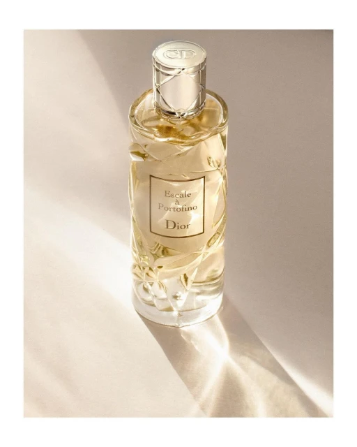 Elegant still life captures a clear glass Dior perfume bottle with a silver cap, labeled 'Escale a Portofino', on a cream background.