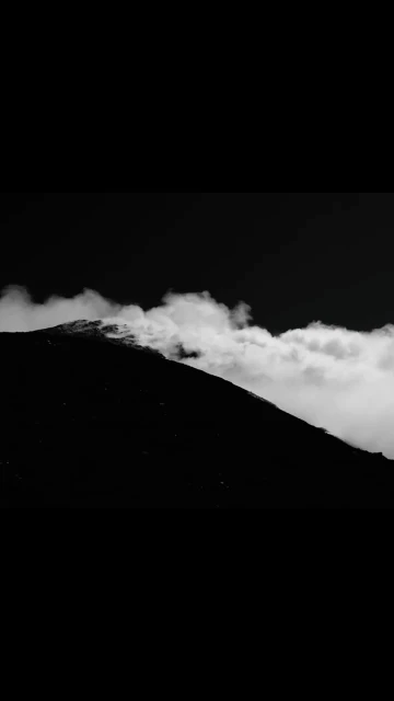 Dramatic black and white landscape of a dark mountain partially obscured by low-lying clouds.