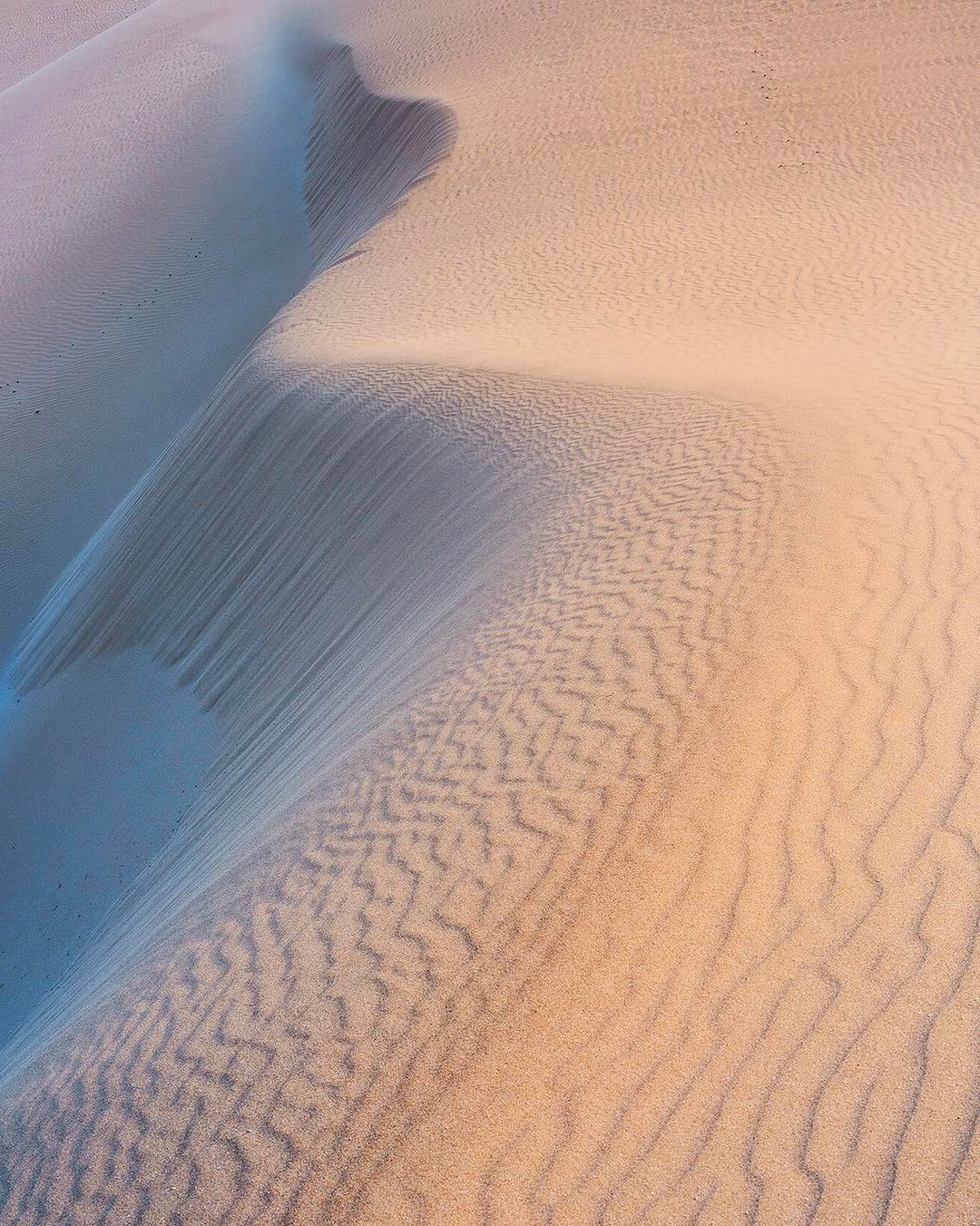 Aerial view of sand dunes featuring patterns and shadows, showcasing the texture and coloration of the desert landscape.