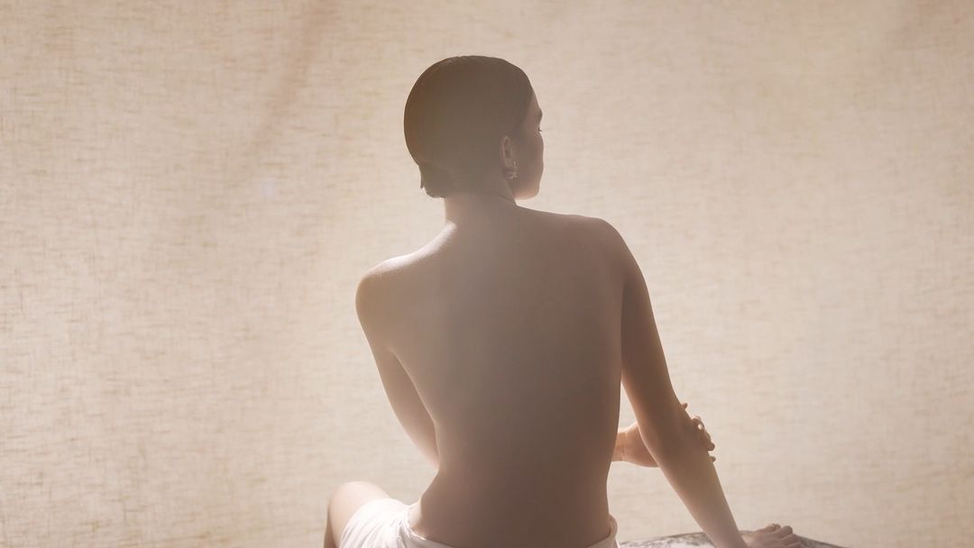 Back view of adult female sitting in diffused light, wearing white towel, tan neutral background.
