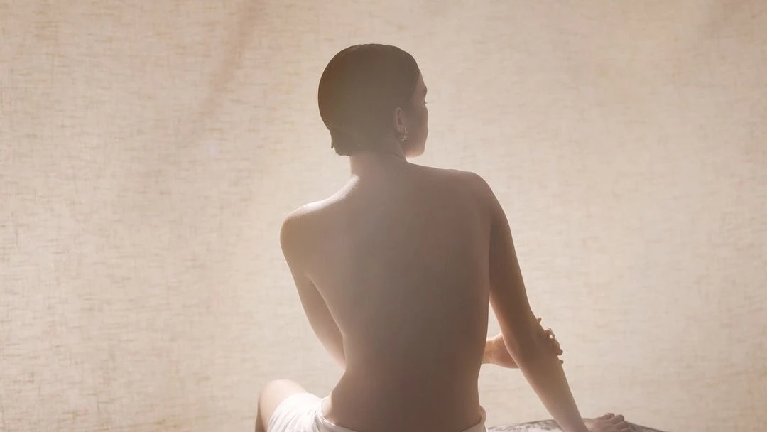 Back view of adult female sitting in diffused light, wearing white towel, tan neutral background.