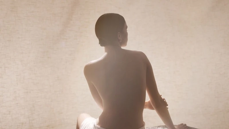 Back view of adult female sitting in diffused light, wearing white towel, tan neutral background.