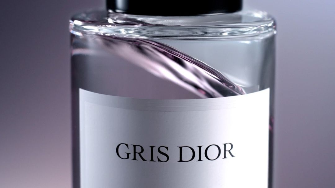 Close-up of a clear glass bottle with a white label reading 'Gris Dior' filled with light gray liquid, set against a gray background.
