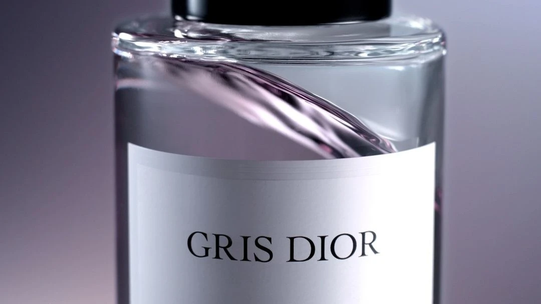 Close-up of a clear glass bottle with a white label reading 'Gris Dior' filled with light gray liquid, set against a gray background.
