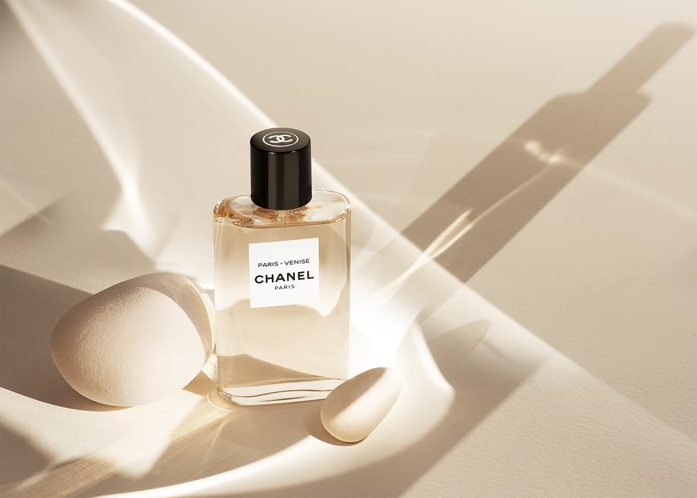 A clear glass bottle of Chanel Paris Venise fragrance sits next to two egg-shaped objects on a cream surface with bright lighting.