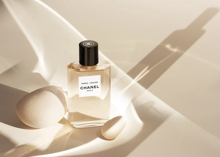 A clear glass bottle of Chanel Paris Venise fragrance sits next to two egg-shaped objects on a cream surface with bright lighting.
