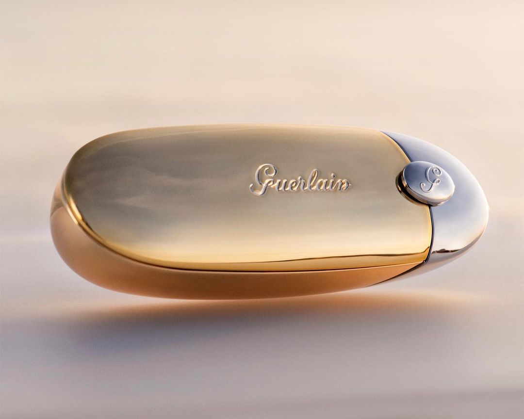 Guerlain lipstick case with gold and silver finish, featuring the brand's logo, showcased on a gradient background for beauty promotion.