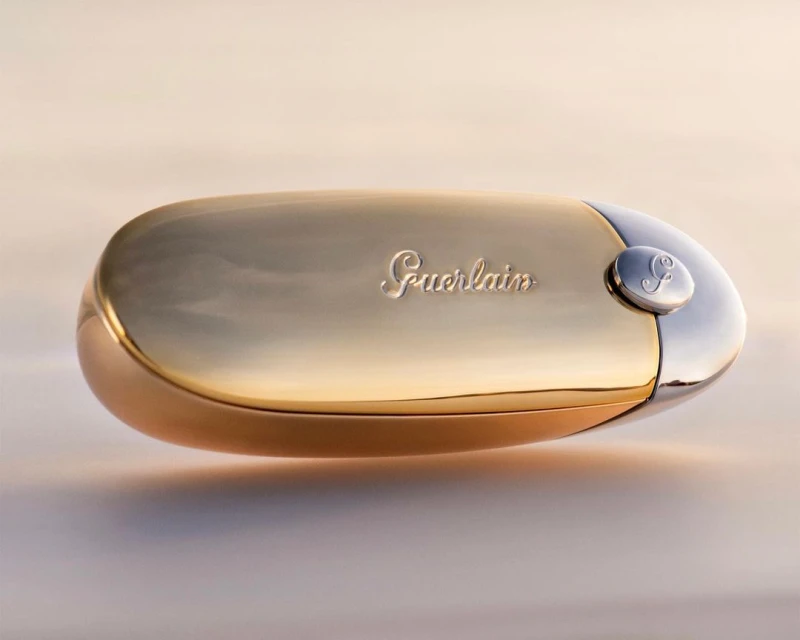 Guerlain lipstick case with gold and silver finish, featuring the brand's logo, showcased on a gradient background for beauty promotion.