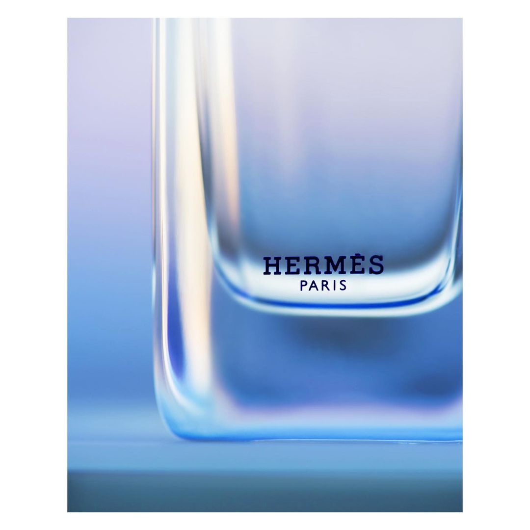 Close-up studio shot of a clear glass Hermes perfume bottle with visible brand logo. Soft blue and white gradient background.