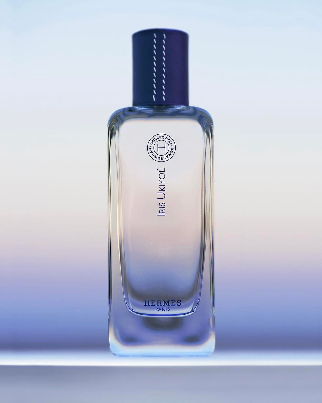 A clear glass perfume bottle with a blue cap stands on a white surface, against a gradient light blue and white background, product shot.