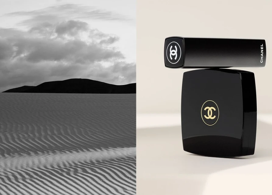 Black Chanel lipstick and compact case stacked in a studio setting against a backdrop of black-and-white landscape with sand dunes.
