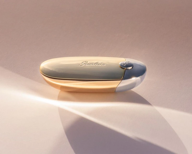 Guerlain KissKiss Bee Glow balm in gold casing, product shot on a pink surface with a curved shadow; makeup, beauty, studio shot.