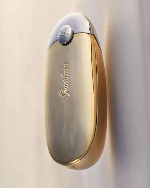 Elegant studio shot of a gold and silver perfume bottle by Guerlain, highlighting its curved shape and reflective surface against a light backdrop.