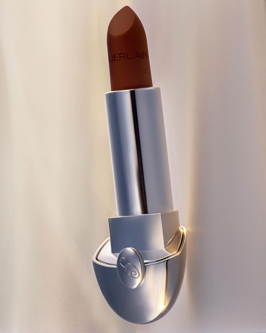 Close-up studio shot of a brown Guerlain lipstick with silver packaging against a beige backdrop with soft lighting.