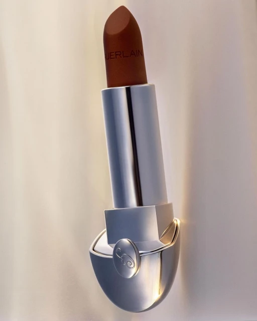 Close-up studio shot of a brown Guerlain lipstick with silver packaging against a beige backdrop with soft lighting.