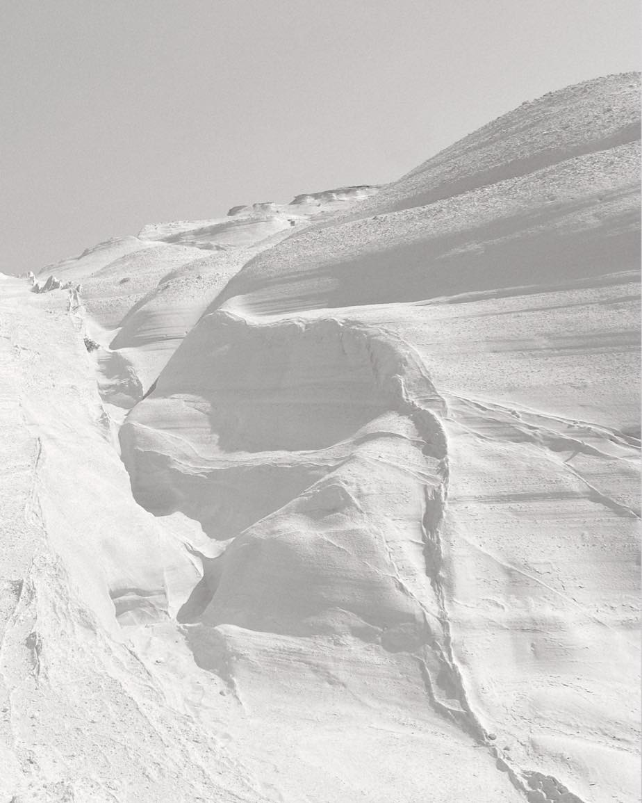 Monochromatic landscape of a snow-covered mountain range against a clear sky, featuring snowdrifts and varied textures.