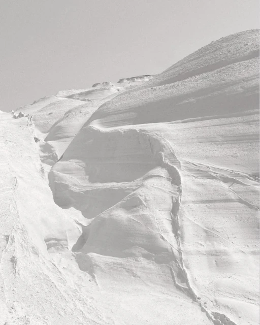 Monochromatic landscape of a snow-covered mountain range against a clear sky, featuring snowdrifts and varied textures.