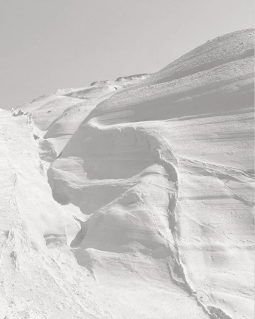 Monochromatic landscape of a snow-covered mountain range against a clear sky, featuring snowdrifts and varied textures.