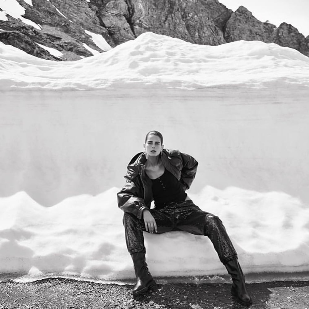 Model seated on a snowbank wearing a dark jacket and trousers against a rocky mountain backdrop in black and white