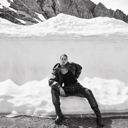 Model seated on a snowbank wearing a dark jacket and trousers against a rocky mountain backdrop in black and white