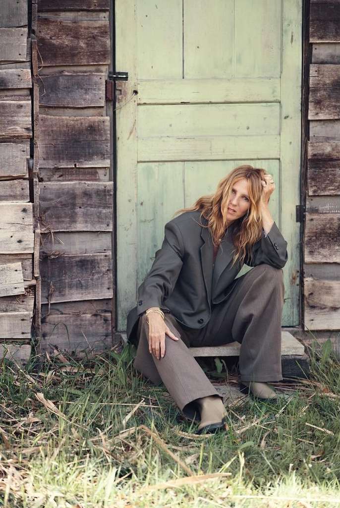 Adult female model sits by a weathered barn door in a casual grey blazer and trousers, in a rural setting with tall grass and sunlight.