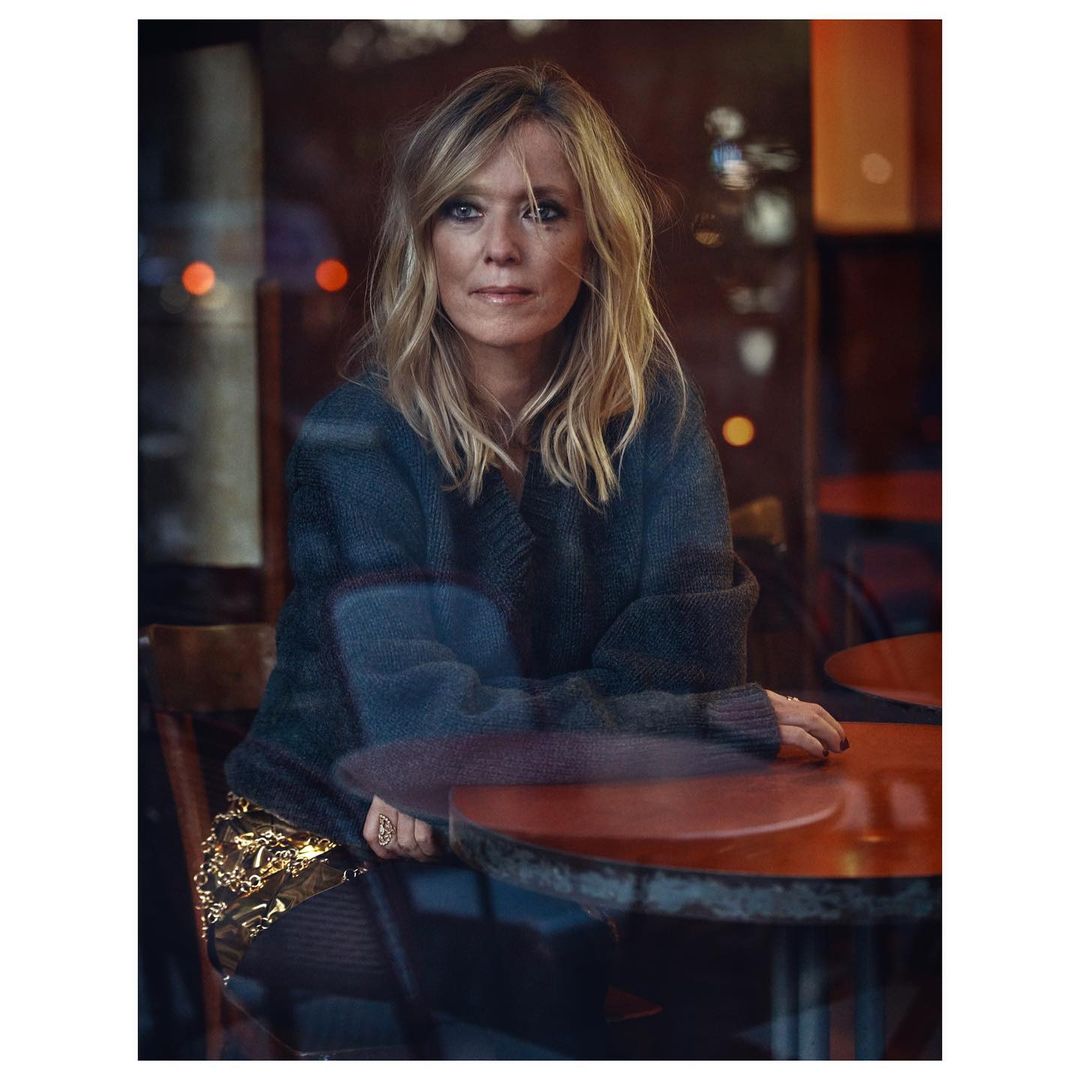 Blonde woman poses indoors by a table. She wears a dark sweater and gold skirt. The photo features soft lighting and reflections.