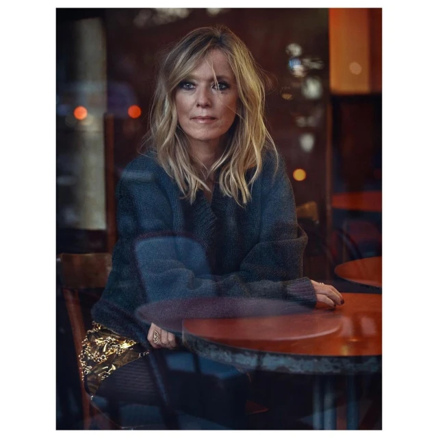 Blonde woman poses indoors by a table. She wears a dark sweater and gold skirt. The photo features soft lighting and reflections.