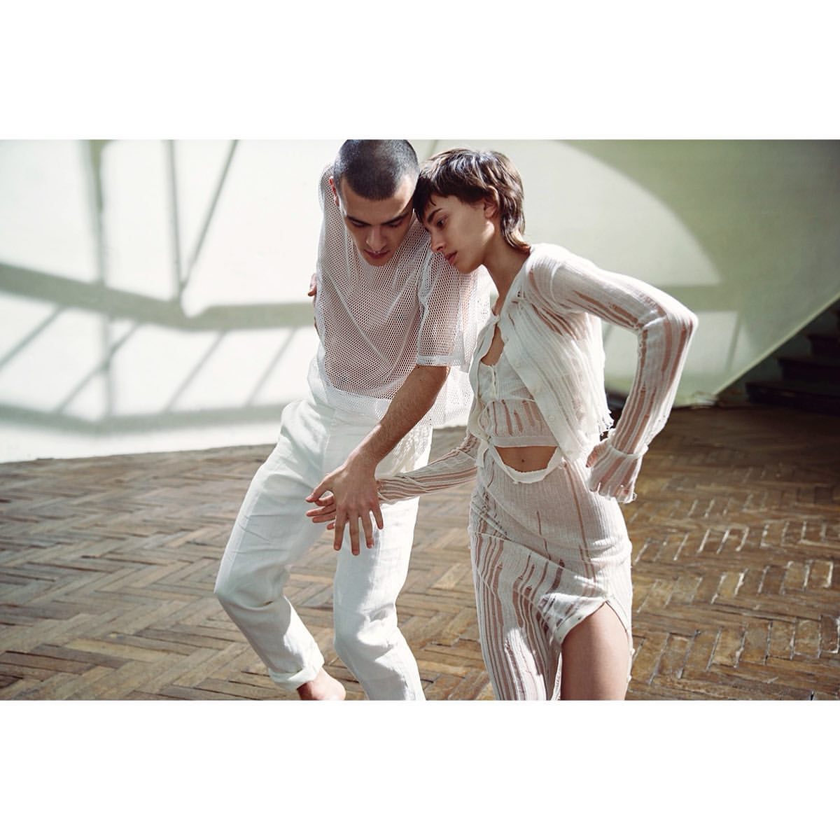 A couple dances in white sheer outfits on a herringbone wood floor, bathed in bright light, creating a modern, minimalist fashion image.
