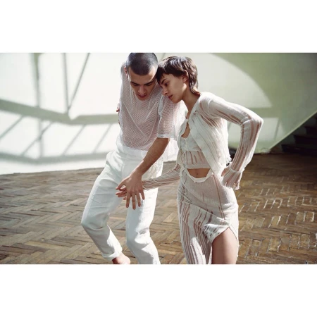 A couple dances in white sheer outfits on a herringbone wood floor, bathed in bright light, creating a modern, minimalist fashion image.