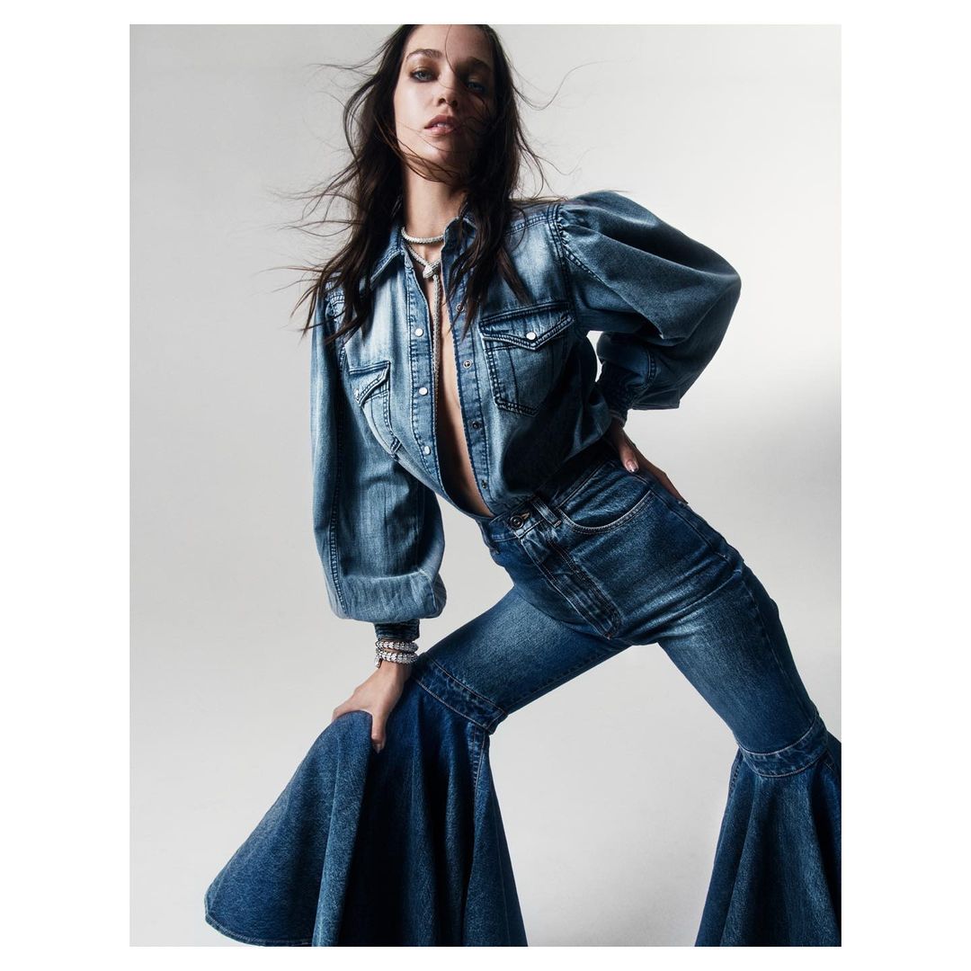 A stylish adult female wearing a denim top and bell-bottom jeans poses indoors in front of a neutral background for a fashion shot.