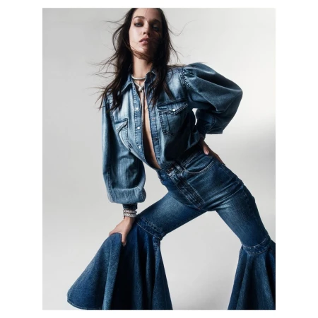 A stylish adult female wearing a denim top and bell-bottom jeans poses indoors in front of a neutral background for a fashion shot.