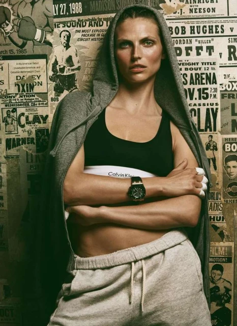Athletic woman in grey sweatsuit poses in front of boxing posters, wearing a Calvin Klein top and watch, with arms crossed.