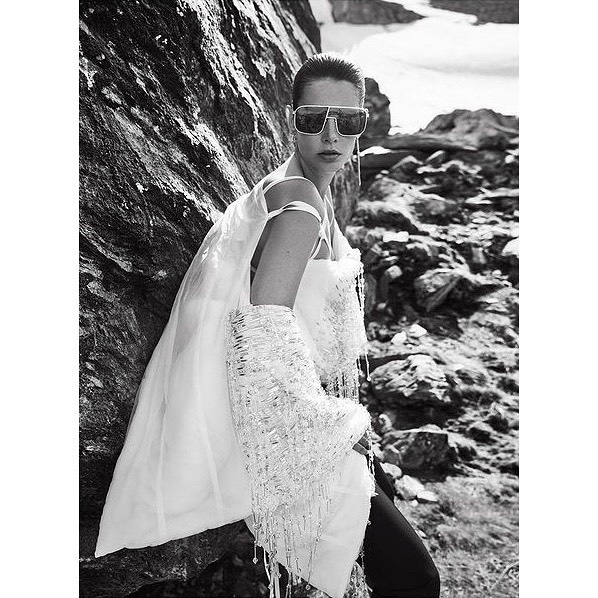 A woman in sheer white top and sunglasses poses next to a rock formation in a monochromatic outdoor fashion shot with an overcast sky.