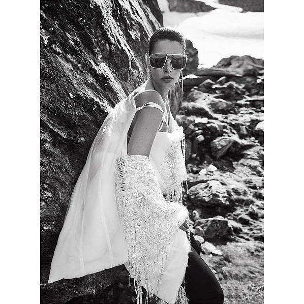 A woman in sheer white top and sunglasses poses next to a rock formation in a monochromatic outdoor fashion shot with an overcast sky.
