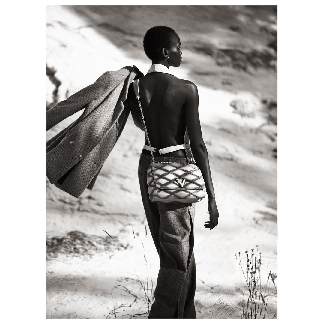Black and white fashion shot of a slender, dark-skinned woman with short hair walking in nature wearing a designer handbag.