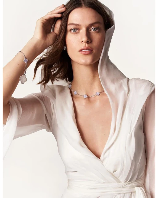 Close-up of a woman wearing a white hooded dress and silver floral jewelry, including a necklace and bracelet, against a plain background.