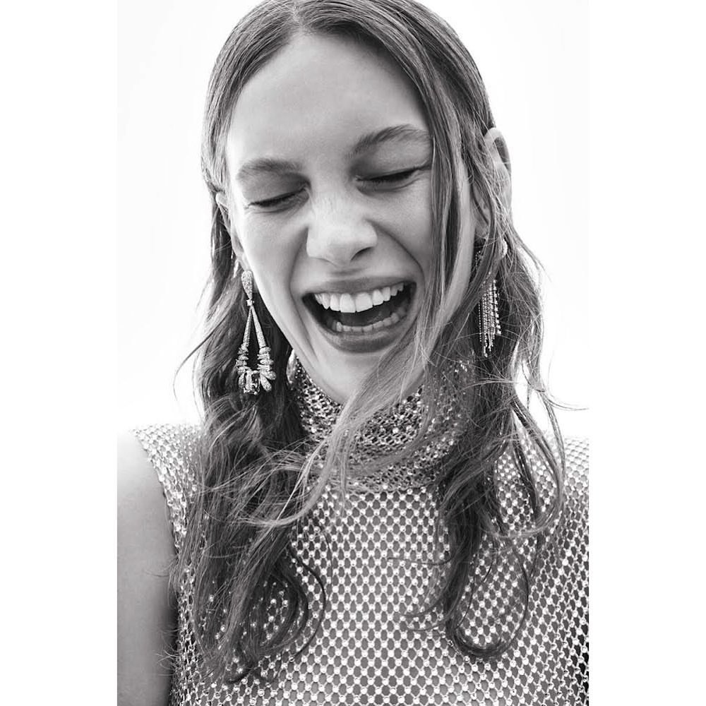 Close-up black and white portrait of a smiling young woman with long wavy hair, wearing a metallic top and dangling earrings.